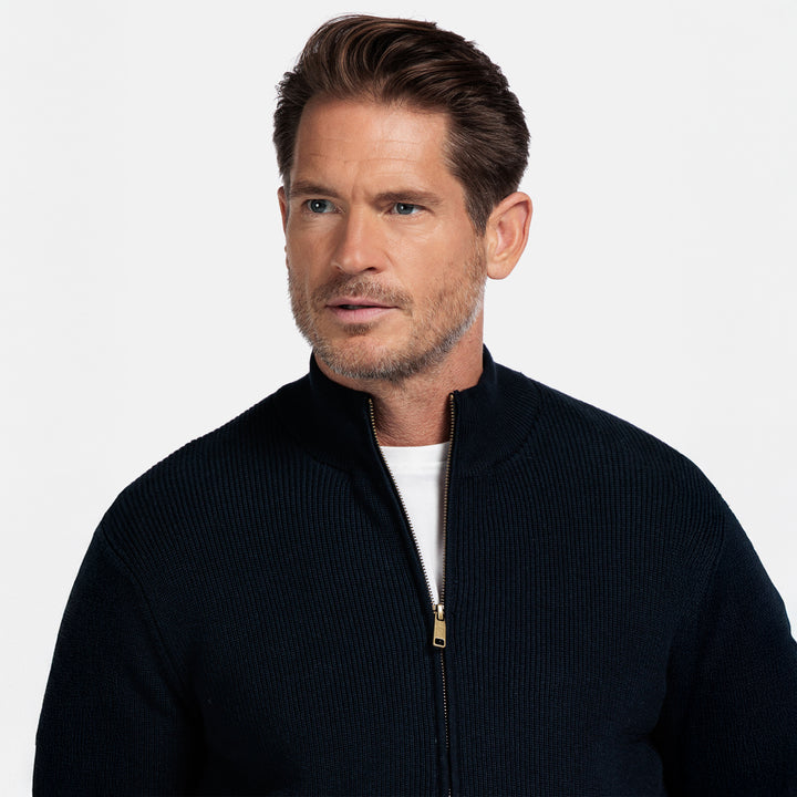 Man wearing a dark blue zip-up sweater over a white shirt on a light gray background