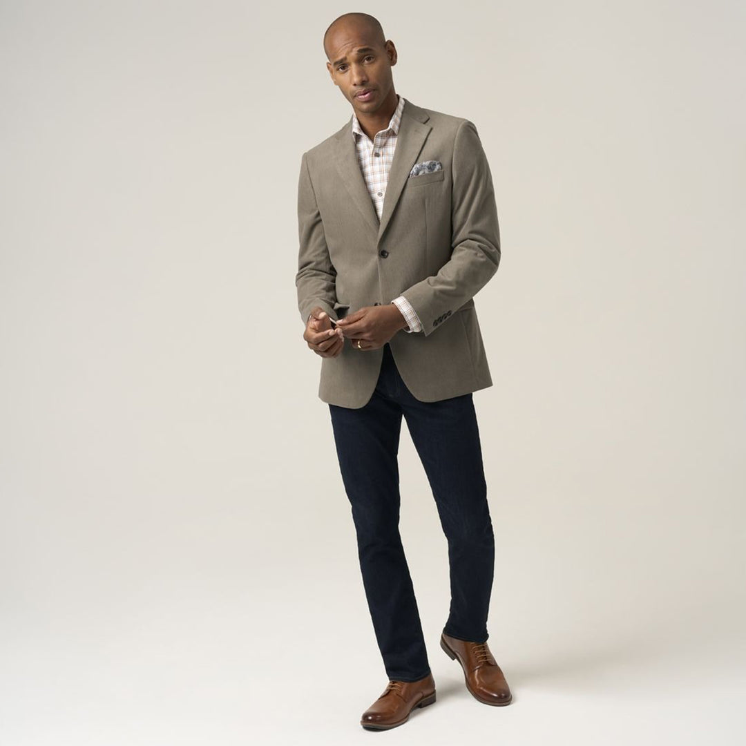 Man wearing a beige blazer over a patterned shirt with dark pants and brown shoes on a white background