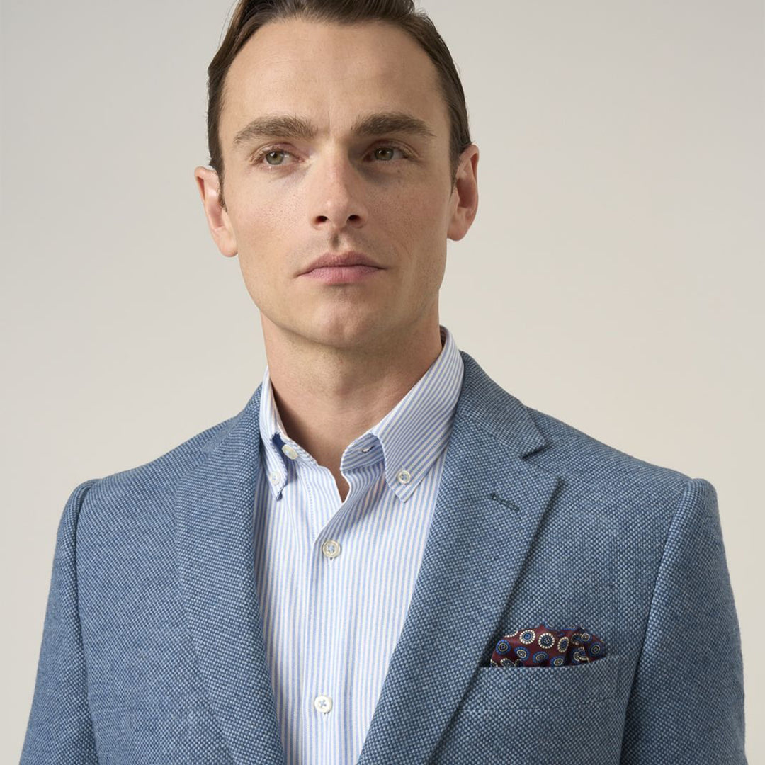 Man wearing a light blue blazer over a striped shirt with a patterned pocket square on a plain background