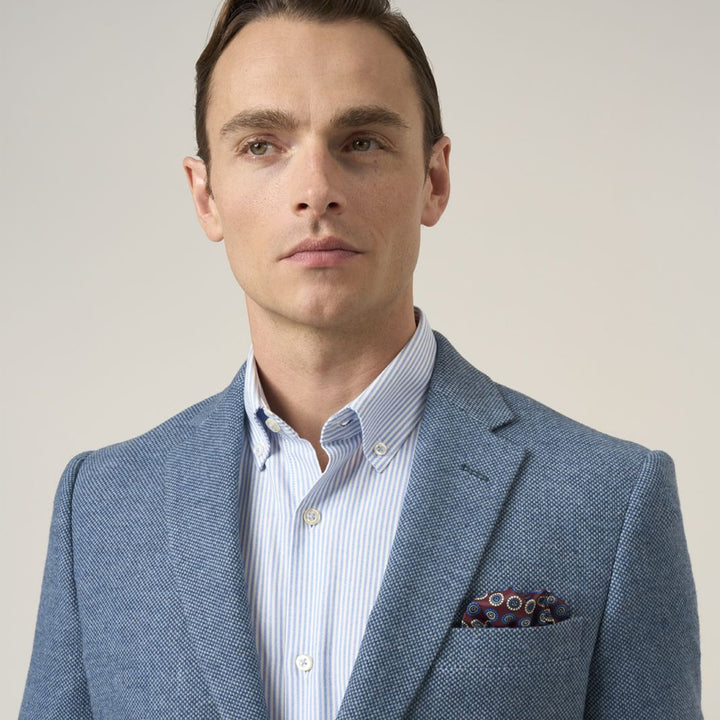 Man wearing a light blue blazer over a striped shirt with a patterned pocket square on a plain background