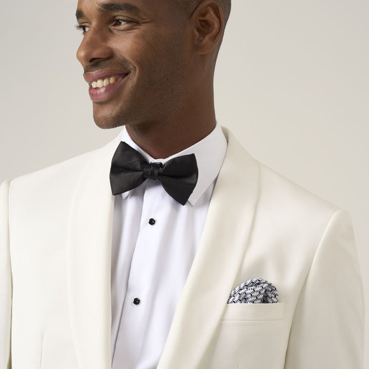 Man wearing a white tuxedo with a black bow tie and pocket square on a light gray background