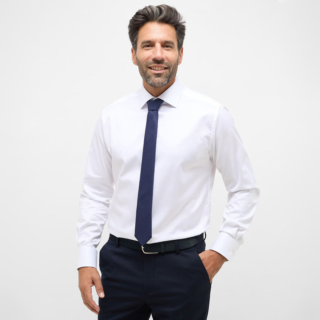 Man wearing a white dress shirt with a navy tie and dark pants on a light gray background