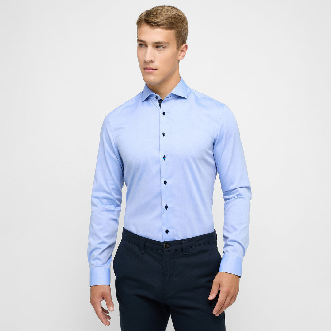 Man wearing a light blue dress shirt and dark pants on a plain background