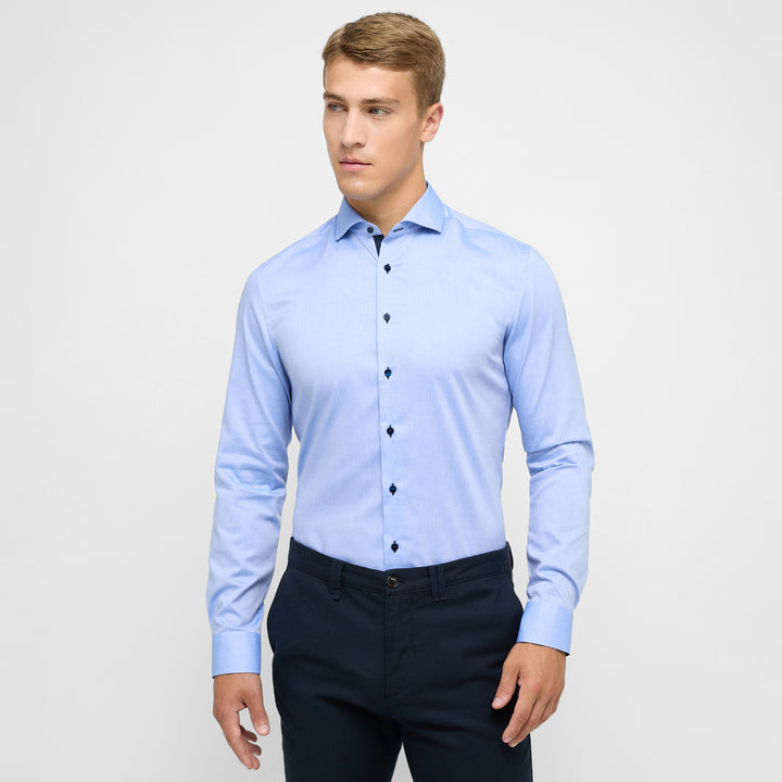 Man wearing a light blue dress shirt and dark pants on a plain background