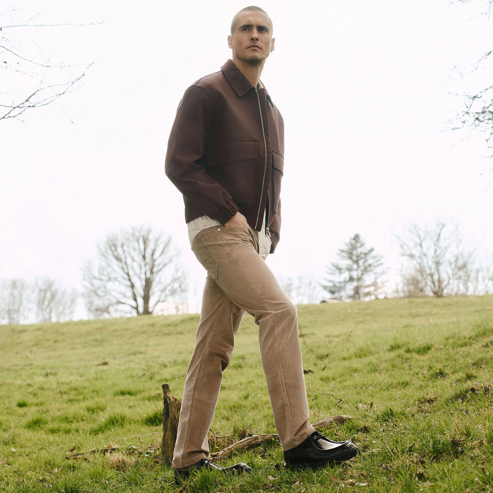 Man in a brown jacket and beige pants standing on a grassy hill with trees in the background