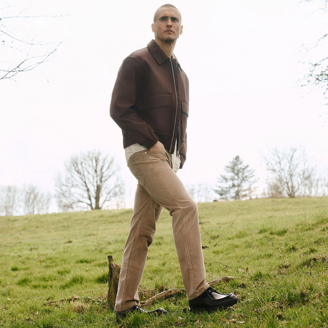 Man in a brown jacket and beige pants standing on a grassy hill with trees in the background
